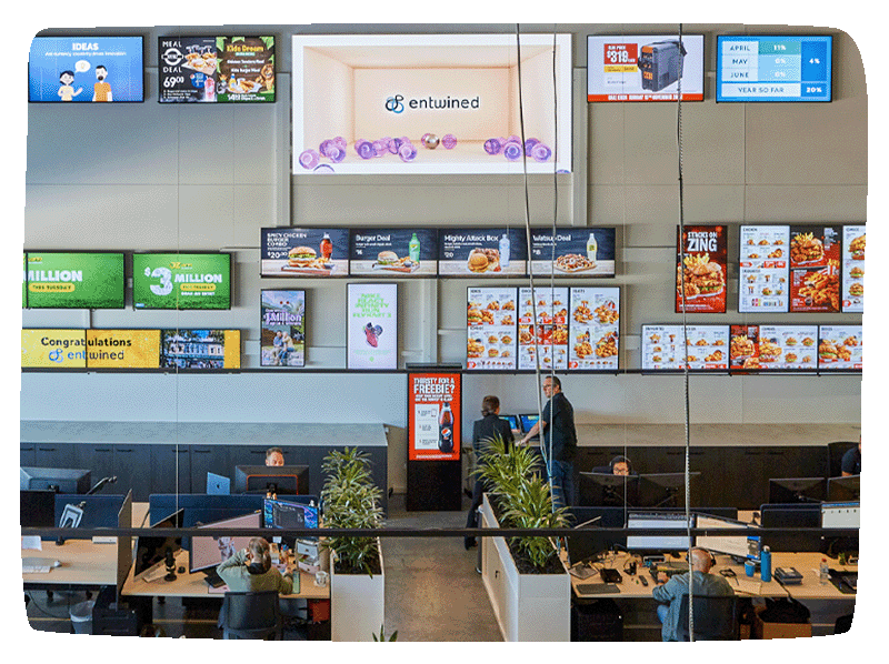 Entwind's office with a large back wall with 15 digital screens on it displaying different content. Two workers standing and talking while two more are at their desk working. 