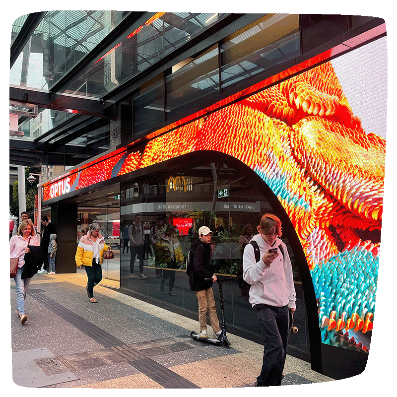 Outside of an Optus building with pedestrians walking past and a large LED screen wrapped around the front.