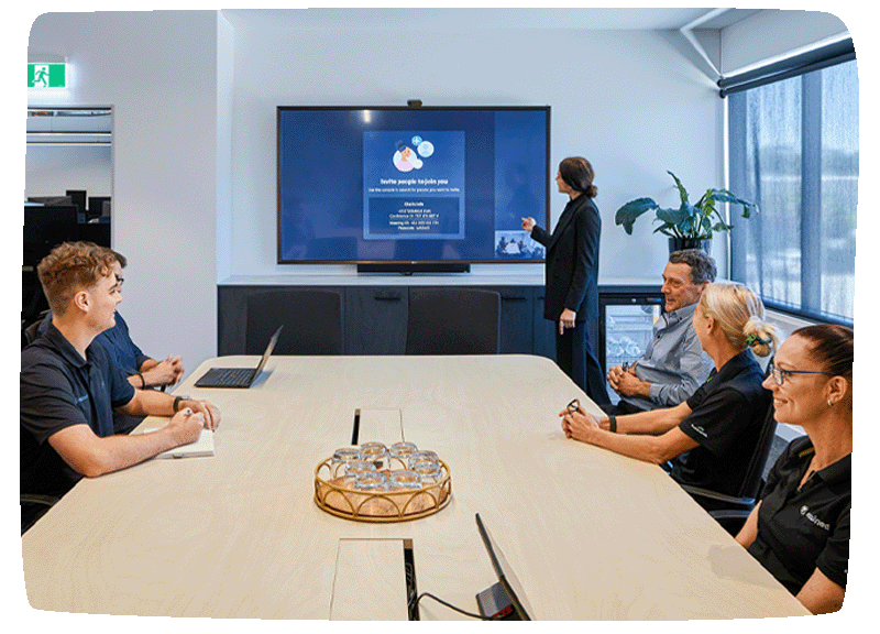 A boardroom filled with employees, while one is presenting a slideshow on the back wall screen.