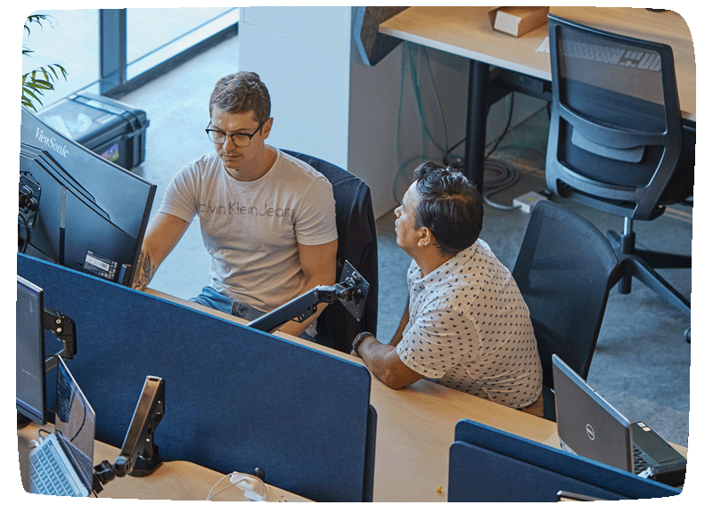 Two people sitting at a desk, one facing the other while the other works on a computer screen.