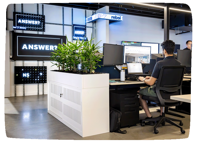 A person sitting at his desk working on duel screens, to the left of him are various digital screens displaying rotating content. 