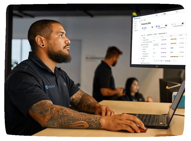 Entwined staff sitting at his desk looking at his monitor. with MyEntwined Dashboard on screen. Two people behind him talking to each other while looking at another screen.