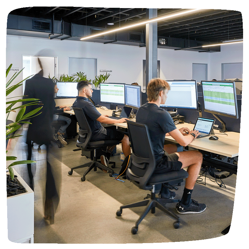 Three workers sitting at their desk in a row, working on their laptops with someone walking behind them.