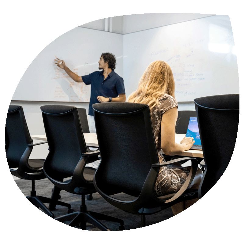 A person writing on a whiteboard while a lady looks on taking notes on her laptop.