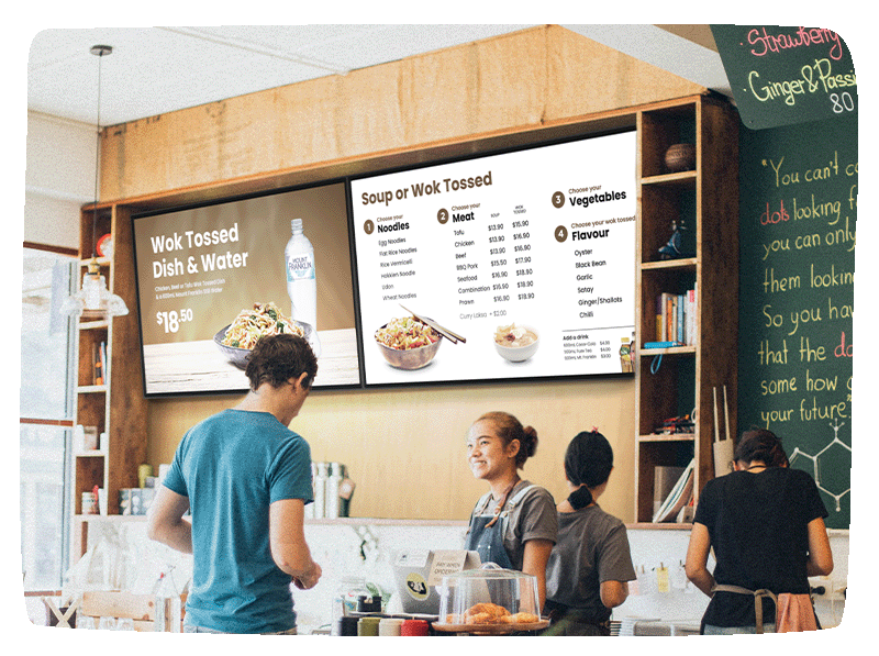 A person ordering at a cafe with someone taking their order at the font desk. Behind her is two digital screens displaying their menu.